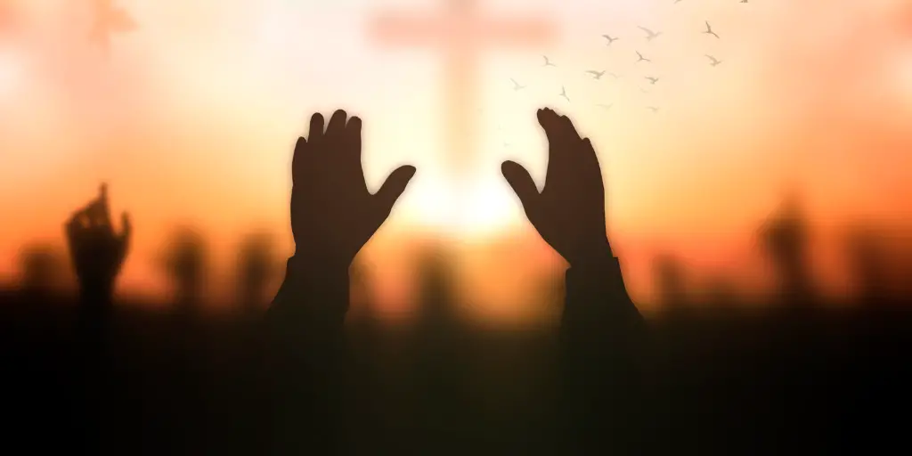 Hands raised to the sky in prayer with the feint image of a cross in the background.