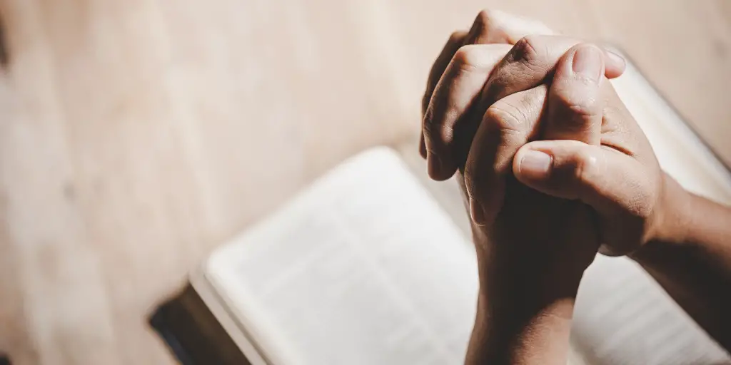 Man clasps his hands in prayer above an open bible.