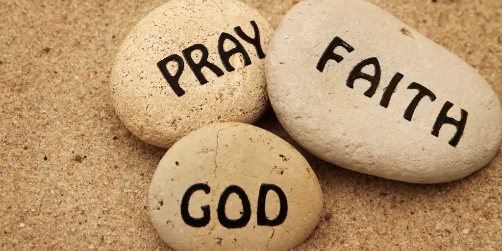 3 rocks with text on them sit next to each other in the sand, they read "Pray," "God," and "Faith"