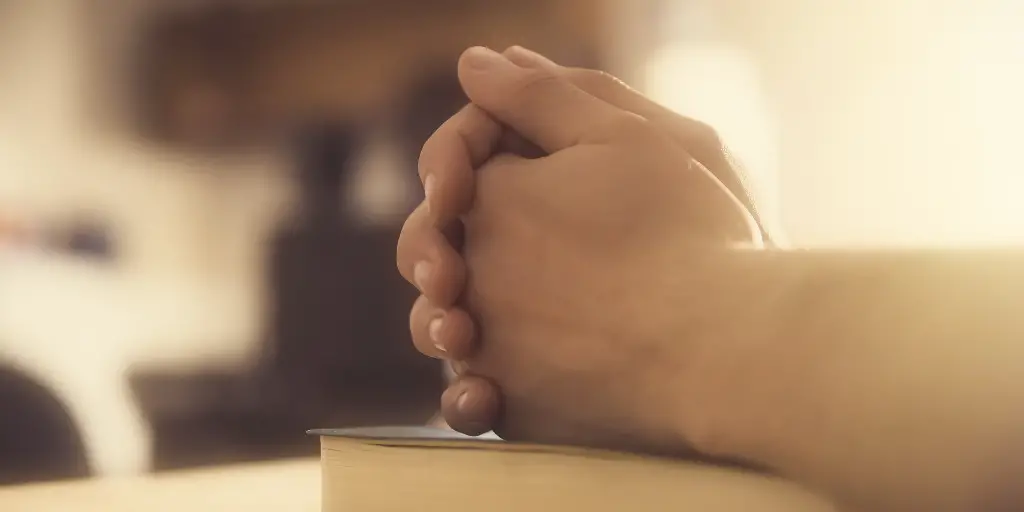 A close-up of a man with his hands clasped a top a Bible, presumably in prayer.