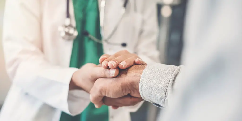 A doctor comforts a patient by holding the patients hand within their own hands.