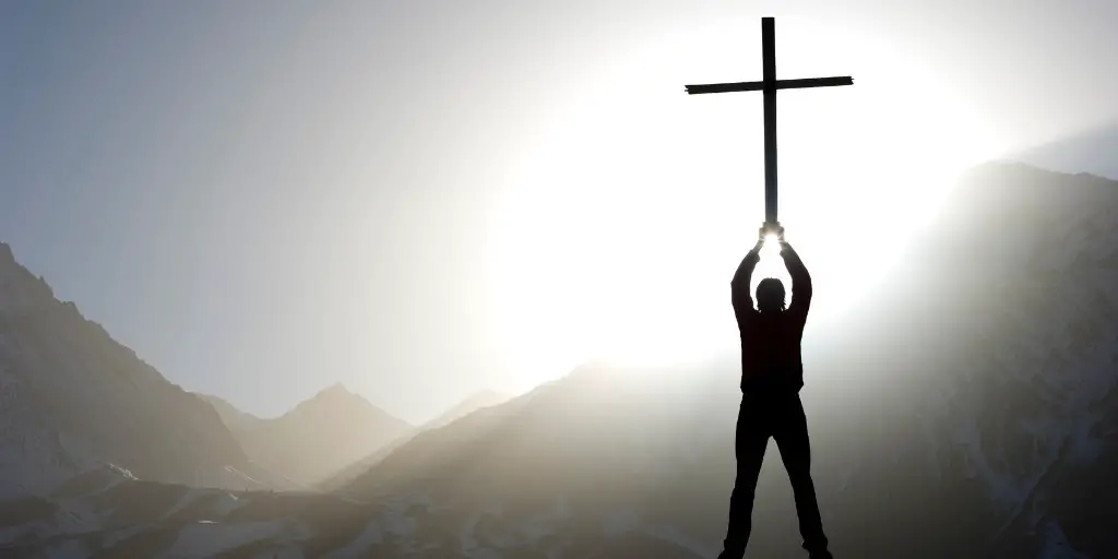 The silhouette of a man as he holds up a giant cross above his head looking into the sun.